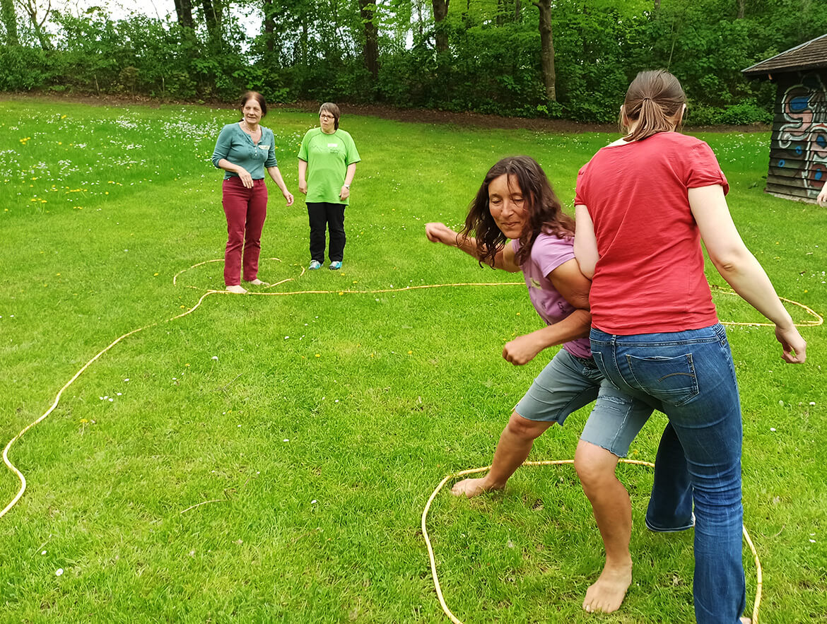 Mit Seilen sind auf einer grünen Wiese Spielbereiche abgesteckt, in denen Menschen unterschiedlichen Alters spielen