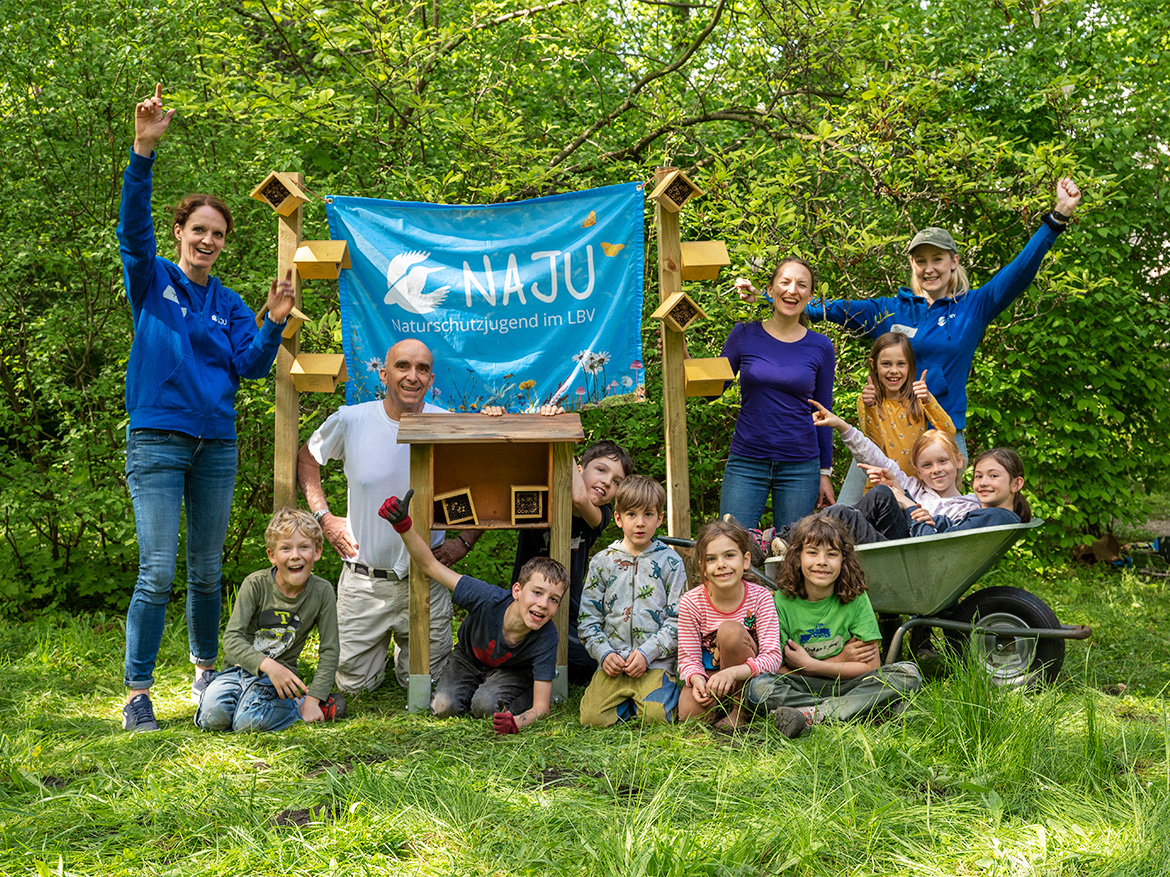 Freudig präsentieren eine Gruppe Kinder mit ein paar Erwachsenen ein Insektenhotel samt NAJU-Fahne