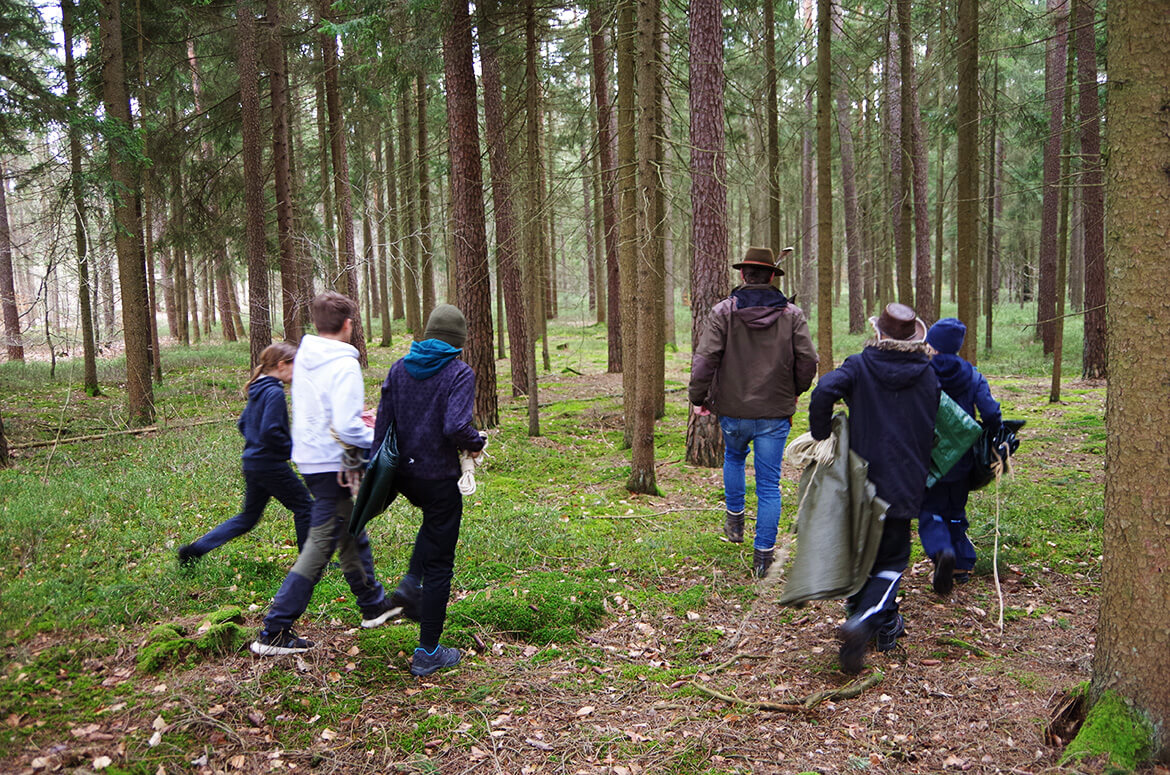 Fünf mit Planen und Seilen bepackte Kinder folgen aufgeregt einem Waldführer mit braunem Hut