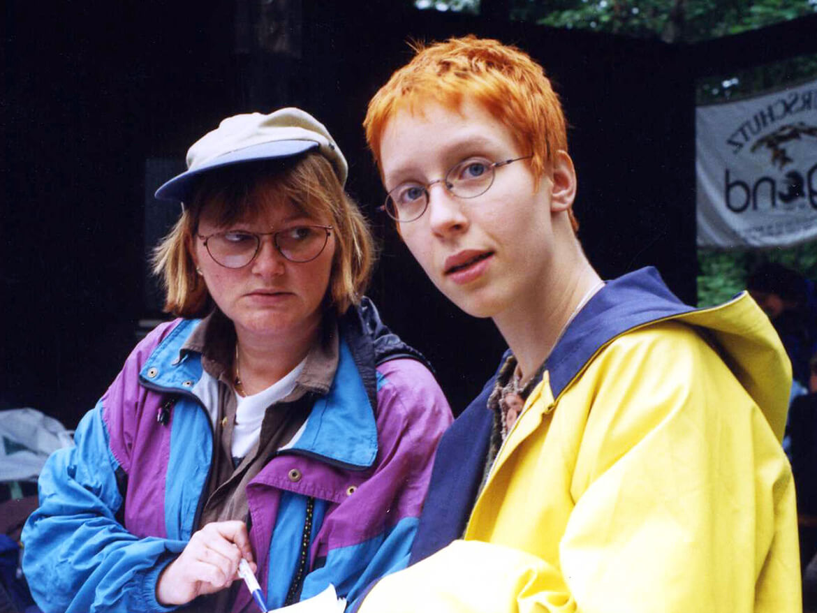 Eine Junge Stefanie mit kurzen, feuerroten Haaren, runder Brille und einem knallgelben Regenmantel steht neben einer ihrer Freundinnen